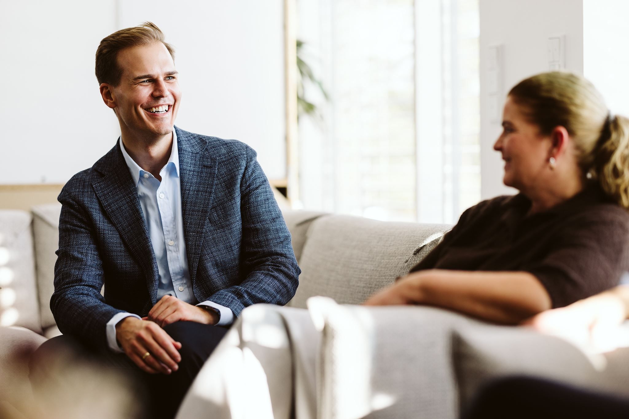 Smilende mann i blazer og kvinne sitter i en sofa og snakker sammen i en uformell samtale