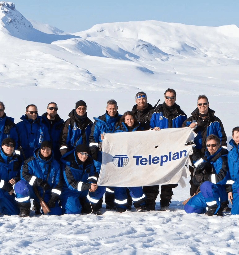Gruppe i blå arbeidsdresser og solbriller poserer med et Teleplan-banner i et snødekket fjellandskap