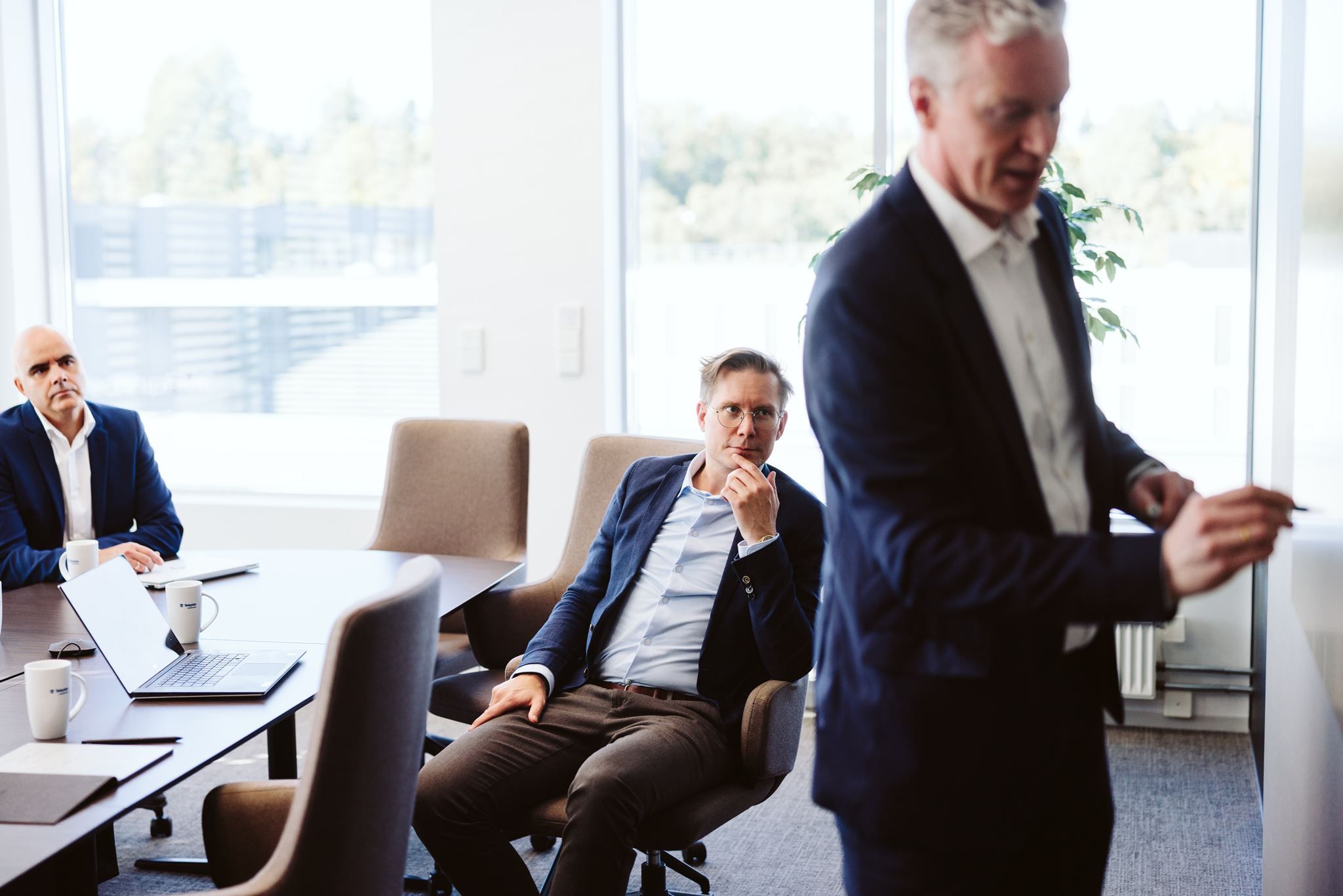 Mann i dress står og presenterer i et møterom mens to kolleger sitter ved bordet og lytter
