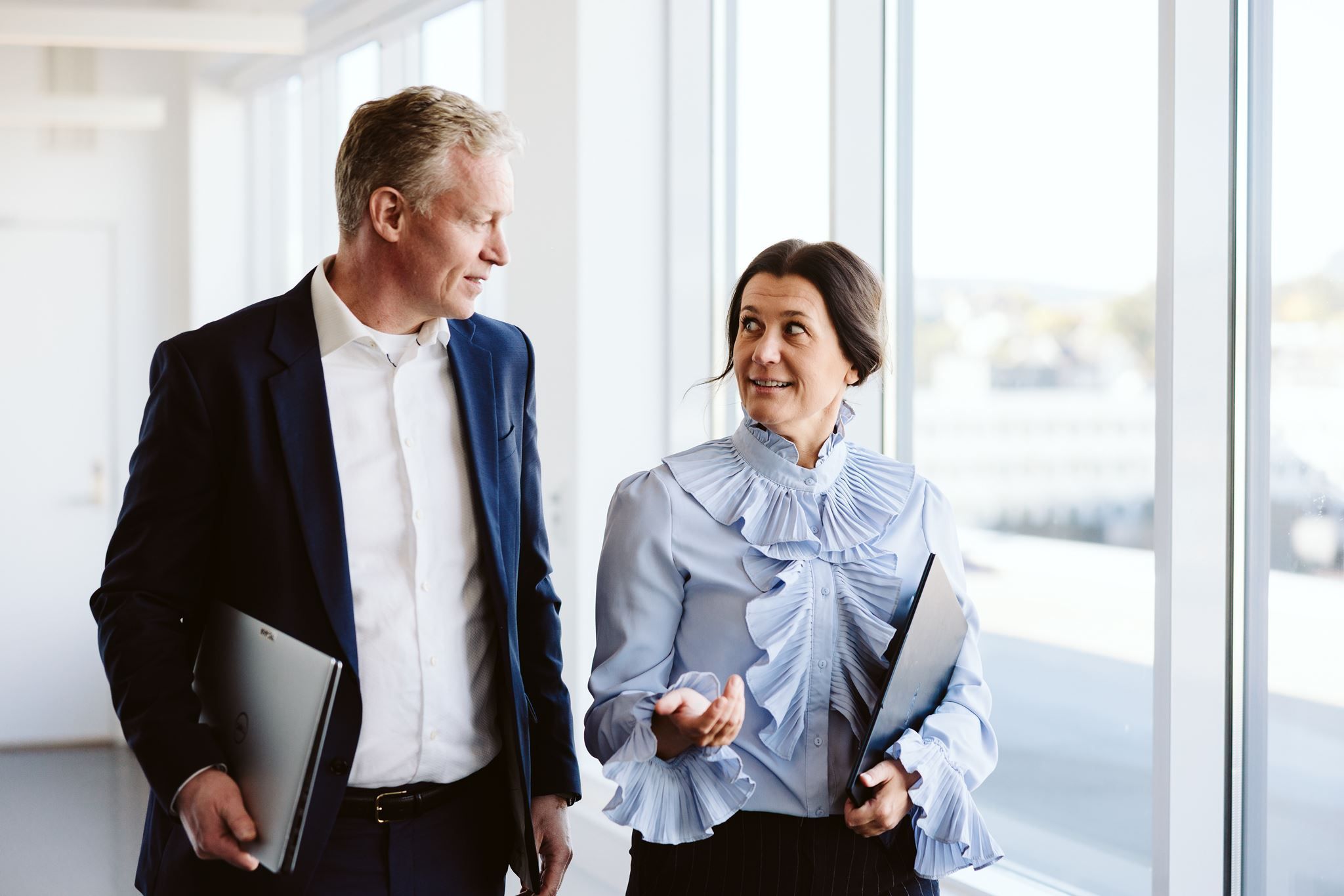 Mann i dress og kvinne i lyseblå bluse går sammen gjennom et lyst kontor med mapper i hånden og snakker